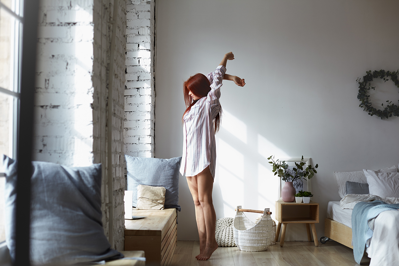 Full lenght shot of attractive young redhead female in long striped shirt standing barefooted on her toes in bedroom and stretching body, raising arms, facing big window, enjoying warm spring sunlight
